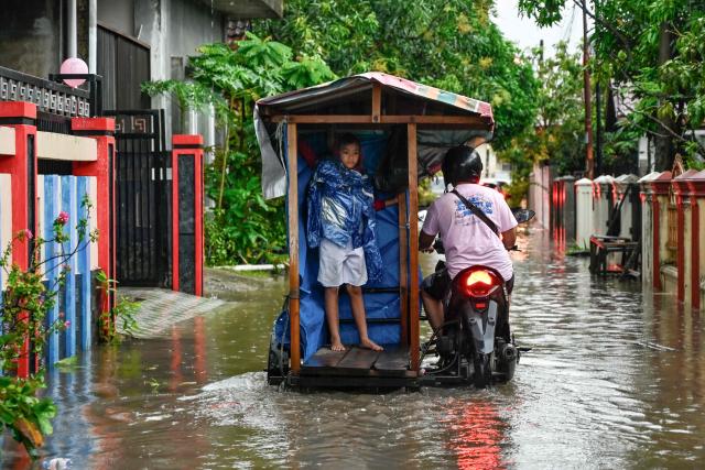 A man transports a child through the floodwaters on an inundated street following heavy rain at a residential area of Darul Imarah on the outskirts of Banda Aceh on November 27, 2025. Tropical storms and monsoon rains have pummelled Southeast and South Asia this month, triggering landslides and flash floods from the rainforests of Indonesia's western Sumatra island to highland plantations in Sri Lanka. (Photo by Chaideer MAHYUDDIN / AFP)