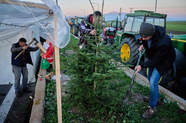 A farmer chops firewood as other farmers plant a christmas tree as they block a national highway with their tractors  on the outskirts of the central Greek city of Larisa on December 9, 2025. Thousands of Greek farmers have since late November blocked highways, mainly in the centre and north of the country, to demand swifter access to EU subsidies delayed by an ongoing probe into multi-million fraud. (Photo by Aggelos NAKKAS / AFP)