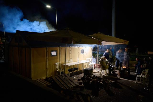 Farmers gather around a fire to warm themselves during a protest blocking a national highway with tractors, on the outskirts of the central Greek city of Larissa on December 9, 2025. Thousands of Greek farmers have since late November blocked highways, mainly in the centre and north of the country, to demand swifter access to EU subsidies delayed by an ongoing probe into multi-million fraud. (Photo by Aggelos NAKKAS / AFP)