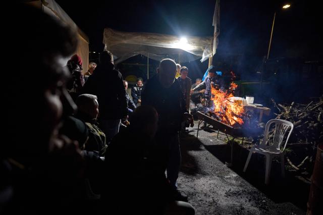 Farmers gather around a fire to warm themselves during a protest blocking a national highway with tractors, on the outskirts of the central Greek city of Larissa on December 9, 2025. Thousands of Greek farmers have since late November blocked highways, mainly in the centre and north of the country, to demand swifter access to EU subsidies delayed by an ongoing probe into multi-million fraud. (Photo by Aggelos NAKKAS / AFP)