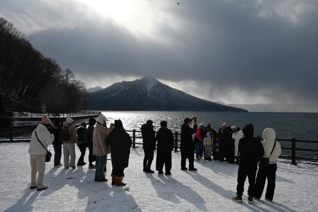 South Korean tourists walk beside Lake Shikotsu, as Mount Fuppushi is seen behind, in the Shikotsu Toya National Park in Japan’s northern Hokkaido prefecture on December 10, 2025. (Photo by GREG BAKER / AFP)