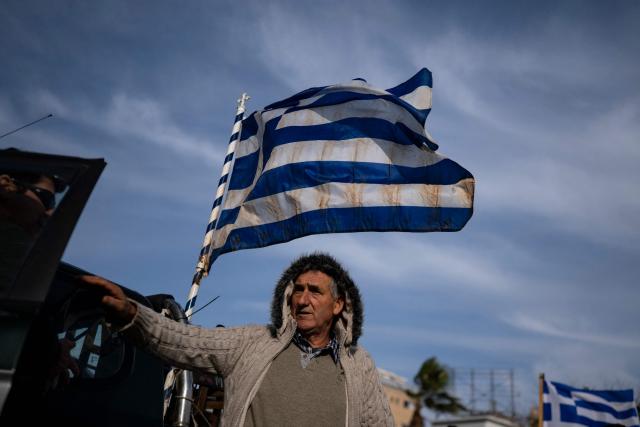A farmer standing beneath a Greek flag looks on as he takes part in the blocking of the passengers' port of the central Greek city of Volos during a demonstration on December 10, 2025. Thousands of Greek farmers have since late November 2025 blocked highways, mainly in the centre and north of the country, to demand swifter access to EU subsidies delayed by an ongoing probe into multi-million fraud. (Photo by Aris MESSINIS / AFP)