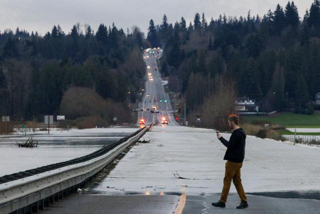 A person stops to capture cellphone images as flood waters from the Snohomish River cover a portion of State Route 9 in Snohomish, Washington, on December 11, 2025. Tens of thousands of people were under evacuation orders Thursday in western North America, after days of heavy rain forced rivers to burst their banks. Storms have battered Washington state in the US and British Columbia over the Canadian border for several days, with rivers continuing to rise. (Photo by Jason Redmond / AFP)