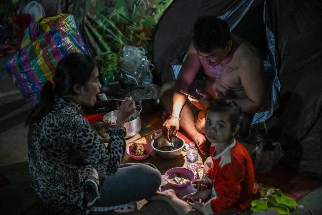 A displaced family uses a flashlight as they eat a meal at a temporary camp in Banteay Meanchey province on December 12, 2025, amid clashes along the Cambodia-Thailand border. (Photo by TANG CHHIN Sothy / AFP)
