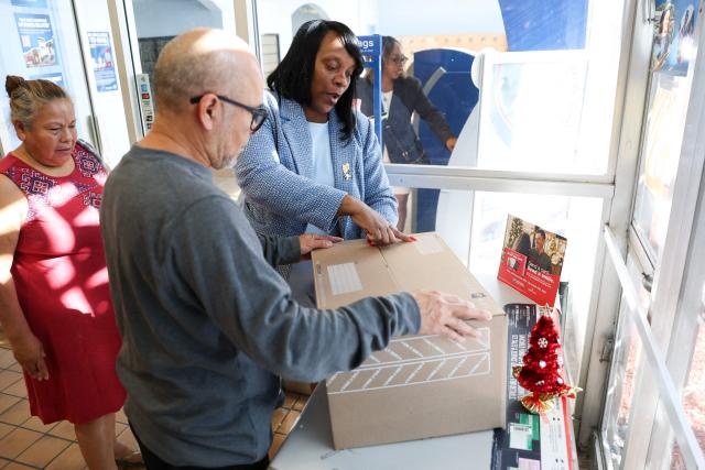 Postmaster Luwanda Smith helps customers with holiday shipping as they mail a package of gifts at the United States Postal Service (USPS) Gardena Post Office in Gardena, California, on December 12, 2025. The USPS reported an increase in expanded daily processing capacity from 60 million to 88 million packages nationwide by deploying more than 600 package sorters, with over 6 billion pieces of mail and packages accepted this holiday season. (Photo by Patrick T. Fallon / AFP)
