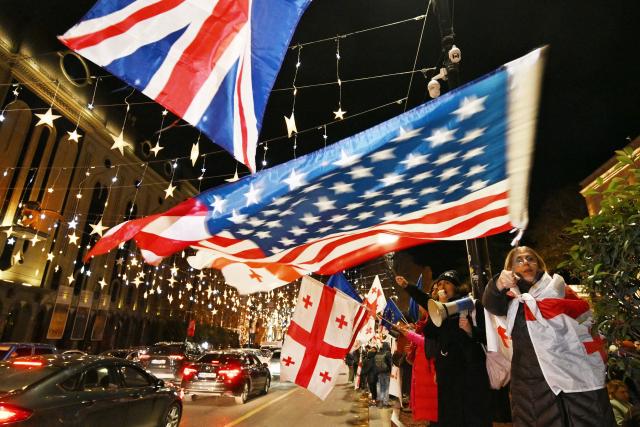 Demonstrators wave Georgian, US and British flags as they protest against the disputed parliamentary elections of 2024 in front of the parliament building in the street decorated with Christmas illuminations in Tbilisi on December 12, 2025. (Photo by Vano SHLAMOV / AFP)