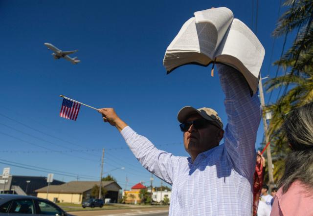 A man waving a US flag and holding a Bible, protests against ICE and Customs and Border Protection near the parking lot of Burlington Coat Factory in Kenner, a suburb of New Orleans, Louisiana, on November 13, 2025. The US Department of Homeland Security announced on December 3 it has launched a federal immigration enforcement operation, named "Operation Catahoula Crunch," in the New Orleans, Louisiana area. (Photo by Matthew Hatcher / AFP)