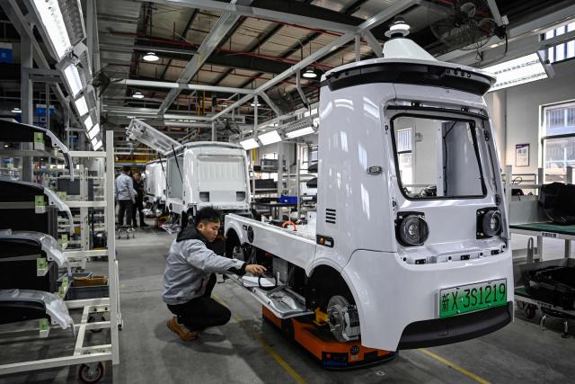 This photo taken on December 4, 2025 shows employees working on a Neolix X3 vehicle production line at a factory of Chinese autonomous delivery vehicle maker Neolix in Yancheng, in eastern China's Jiangsu province. In a light-filled workshop in eastern China, a robotic arm moved a partially assembled autonomous vehicle as workers calibrated its cameras, typical of the incremental automation being adopted even across smaller factories in the world's manufacturing powerhouse. (Photo by Jade GAO / AFP) / To go with 'CHINA-ECONOMY-MANUFACTURING-TECHNOLOGY-AI, FOCUS' by Jing Xuan TENG