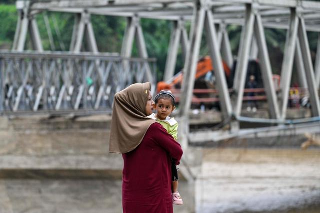 A woman holds her child while waiting to cross a river after a bridge was damaged by flooding in Juli, in Indonesia's Aceh province on December 14, 2025. Devastating floods and landslides have killed 1,006 people in Indonesia, rescuers said December 13 as the Southeast Asian nation grapples with the huge scale of relief efforts. (Photo by CHAIDEER MAHYUDDIN / AFP)