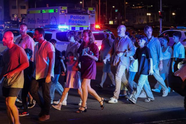 People cross a street next to police vehicles after a shooting incident at Bondi Beach in Sydney on December 14, 2025. Australian police said two people were in custody following reports of multiple gunshots on December 14 at Sydney's famed Bondi Beach, urging the public to take shelter. (Photo by DAVID GRAY / AFP)