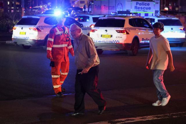 People cross a street next to police vehicles after a shooting incident at Bondi Beach in Sydney on December 14, 2025. Australian police said two people were in custody following reports of multiple gunshots on December 14 at Sydney's famed Bondi Beach, urging the public to take shelter. (Photo by DAVID GRAY / AFP)