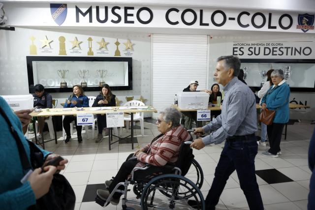 A man pushes a woman in a wheelchair in front of a polling station during the presidential runoff election at Monumental Colo-Colo club Stadium in Santiago, Chile on December 14, 2025. Chileans head to the polls for a presidential runoff between Jeannette Jara, a communist backed by a broad left coalition, and Jose Antonio Kast, a devout far-right politico promising a hard line on security and migration. (Photo by Raul BRAVO / AFP)