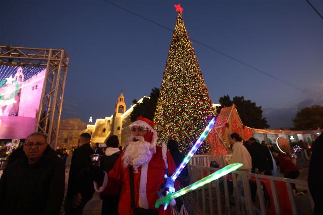 A man dressed as Santa Clause greets pilgrims and Palestinians as he stands in front of a Christmas tree in Manger Square, in the Israeli-occupied West Bank city of Bethlehem on December 14, 2025. Christmas cheer returned to the traditional birthplace of Jesus Christ on December 6, 2025, the first time the city has held the usual celebrations since the outbreak of the war in Gaza following Hamas's attack on Israel in October 2023. (Photo by HAZEM BADER / AFP)