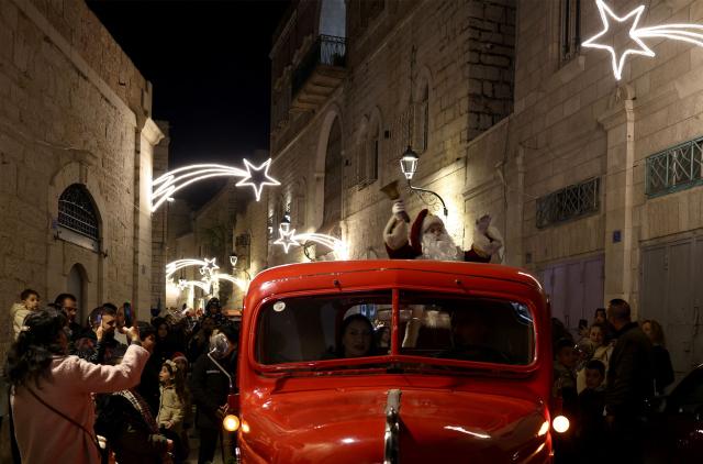 Santa Clause rides in the back of a red truck driving towards Manger Square as Palestinians and pilgrims arrive to visit the Christmas tree, in the Israeli-occupied West Bank city of Bethlehem on December 14, 2025. Christmas cheer returned to the traditional birthplace of Jesus Christ on December 6, 2025, the first time the city has held the usual celebrations since the outbreak of the war in Gaza following Hamas's attack on Israel in October 2023. (Photo by HAZEM BADER / AFP)
