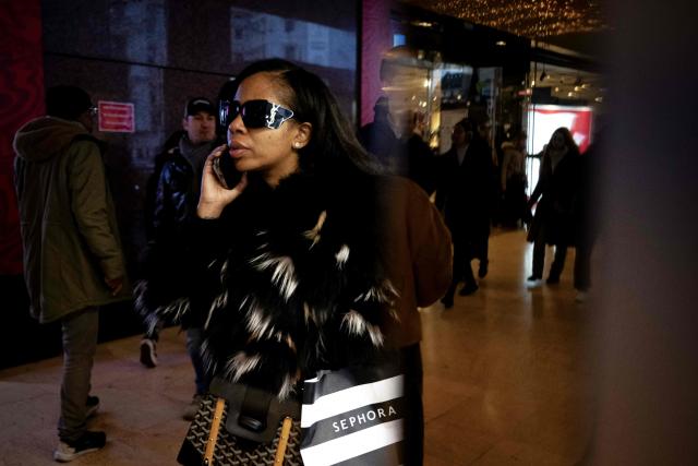 A pedestrian wearing sunglasses uses her phone as she walks past the Galeries Lafayette, days before Christmas, in Paris on December 14, 2025. (Photo by Anna KURTH / AFP)