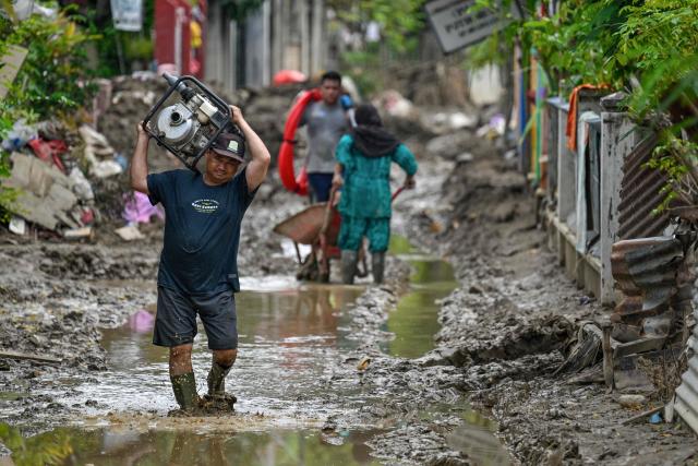 A man wades through the mud as he carries a pump generator to clean houses at a flood-affected area in Meureudu, in Indonesia's Aceh province on December 14, 2025. Devastating floods and landslides have killed 1,006 people in Indonesia, rescuers said December 13 as the Southeast Asian nation grapples with the huge scale of relief efforts. (Photo by CHAIDEER MAHYUDDIN / AFP)