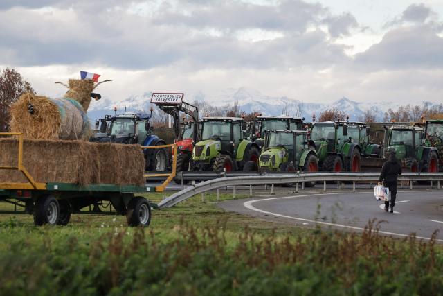 A man walks with water bottles past a line of tractors parked on the A64 motorway as part of the fourth day of a blockade in protest against health measures put in place to eradicate lumpy skin disease affecting cattle, in Carbonne, south-western France, on December 15, 2025. (Photo by Valentine CHAPUIS / AFP)