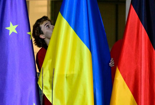 A worker installs the Ukrainian flag between the flags of Europe (L) and Germany (R) prior to a press conference of the German Chancellor and Ukraine's President at the Chancellery in Berlin, Germany, on December 15, 2025. Ukrainian President Volodymyr Zelensky travelled to Berlin to meet European leaders as well as US President Donald Trump's envoy, heads of the EU and of the NATO for talks on how to end the grinding war with Russia. (Photo by John MACDOUGALL / AFP)