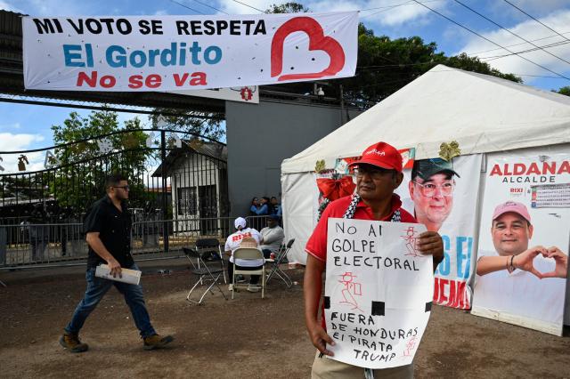 A supporter of the ruling Libertad y Refundacion (LIBRE) party holds a sign reading "No to the electoral coup and pirate Trump out of Honduras" near the National Institute for Professional Training (INFOP), where the National Electoral Council (CNE) is counting 2,800 ballots with inconsistencies from the general elections, in Tegucigalpa, on December 15, 2025. LIBRE called for protests in the streets to denounce electoral fraud and the interference of US President Donald Trump in the elections. Today marks two weeks since Hondurans have been waiting to find out who their next president will be, as they await a special recount that will determine the winner between conservative Nasry Asfura, supported by Donald Trump, and right-wing Salvador Nasralla. (Photo by Orlando SIERRA / AFP)