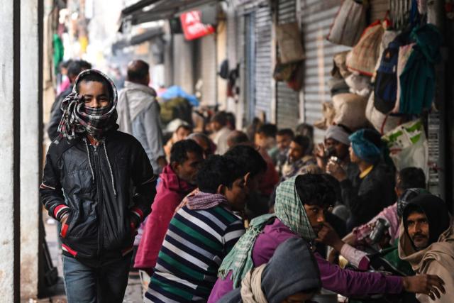 A child wearing a winter stole walks past daily wage workers at a pavement in the old quaters of Delhi on December 16, 2025. (Photo by Arun SANKAR / AFP)