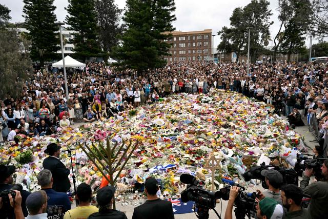 Mourners stand near tributes piled together in memory of the victims of a shooting at Bondi Beach, in Sydney on December 16, 2025. Australia's leaders have agreed to toughen gun laws after attackers killed 15 people at a Jewish festival on Bondi Beach, the worst mass shooting in decades decried as antisemitic "terrorism" by authorities. (Photo by Saeed KHAN / AFP)