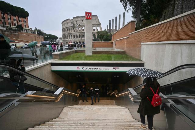 A pedestrian with an umbrella enters the 'Colosseo' new Metro station, designed as a museum to showcase the archaeological findings uncovered during its construction, as it opens to the public, with the Colosseum in the background, in Rome on December 16, 2025. (Photo by Filippo MONTEFORTE / AFP)