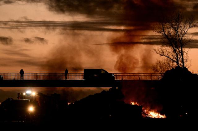 TOPSHOT - People look down from an overpass as a fire burns at a farmers blockade of the A63 motorway at the Cestas interchange in Gironde, south-western France on December 16, 2025. French farmers have been protesting against the government's mandatory culling protocol for cattle herds affected by lumpy skin disease (dermatose nodulaire contagieuse), a viral disease first detected in France in June 2025 that has led to the slaughter of over 3,000 cattle across more than 110 outbreaks nationwide. (Photo by Philippe LOPEZ / AFP)