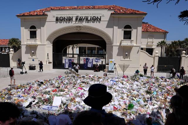 Mourners stand near tributes piled together at the front of the Bondi Pavilion, in memory of the victims of the Bondi Beach shooting, in Sydney on December 17, 2025. Australia held the first funeral on December 17 for victims of the Bondi Beach mass shooting, as large crowds gathered to grieve a rabbi slain in the attack. Sajid Akram and his son Naveed opened fire on a Jewish festival at the famed surf beach on December 14 evening, killing 15 people and wounding dozens more. (Photo by DAVID GRAY / AFP)