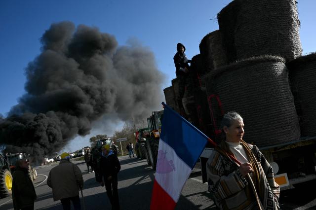 A woman holds a French National flag as farmers from the Jeunes Agriculteurs (JA, Young Farmers), Confederation Paysanne, and  Coordination Rurale (CR) unions block a roundabout with tractors during a protest against the mass slaughter of cows to control the infectious bovine disease nodular dermatitis, also known as lumpy skin disease, in Carcassonne, southern France, on December 17, 2025. (Photo by Matthieu RONDEL / AFP)