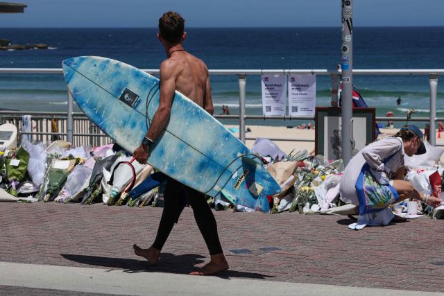 A surfer walks past floral tributes left at the promenade of Bondi Beach in Sydney on December 18, 2025, to honour victims of the shooting that took place there on December 14. The attack at Bondi Beach on December 14 was one of the deadliest in Australian history. (Photo by DAVID GRAY / AFP)