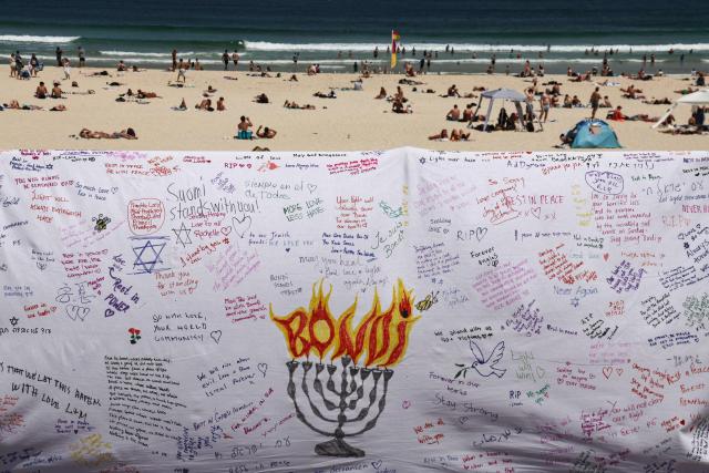 Beachgoers are seen behind a piece of cloth bearing messages left by mourners at the promenade of Bondi Beach in Sydney on December 18, 2025, to honour victims of the shooting that took place there on December 14. The attack at Bondi Beach on December 14 was one of the deadliest in Australian history. (Photo by DAVID GRAY / AFP)