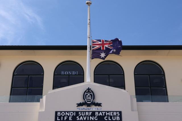 Australia's flag flies at half-mast at the Bondi Surf Bathers Life Saving Club near the promenade of Bondi Beach, where mourners have left floral tributes to honour victims of the shooting that took place there on December 14, in Sydney on December 18, 2025. The attack at Bondi Beach on December 14 was one of the deadliest in Australian history. (Photo by DAVID GRAY / AFP)