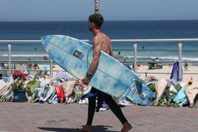 A surfer walks past floral tributes left at the promenade of Bondi Beach in Sydney on December 18, 2025, to honour victims of the shooting that took place there on December 14. The attack at Bondi Beach on December 14 was one of the deadliest in Australian history. (Photo by DAVID GRAY / AFP)