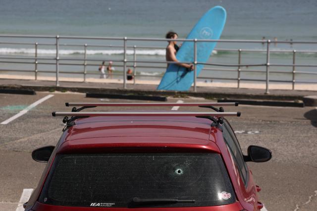 A surfer walks past a car whose rear windshield has a bullet hole in it, at the promenade of Bondi Beach in Sydney on December 18, 2025, after the shooting that took place there on December 14. The attack at Bondi Beach on December 14 was one of the deadliest in Australian history. (Photo by DAVID GRAY / AFP)