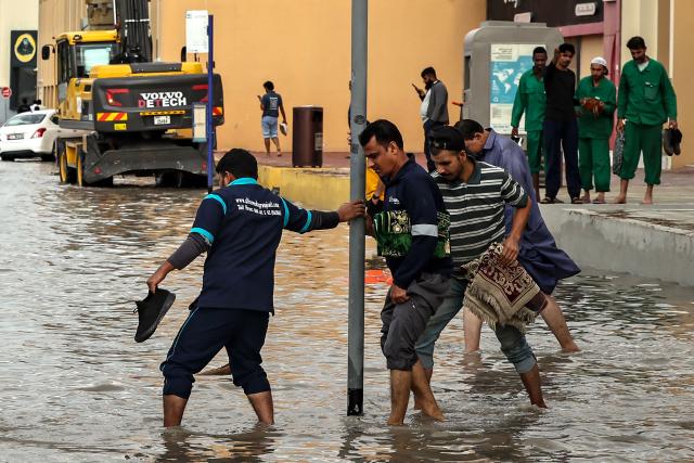 Men hold up their trousers legs to wade through flood waters as they cross the road following heavy rains, in Dubai on December 19, 2025. Dubai police urged residents on December 18, 2025, to stay indoors unless it was "absolutely necessary" as heavy rainfall was expected in the desert country which saw record downpours last year. (Photo by FADEL SENNA / AFP)