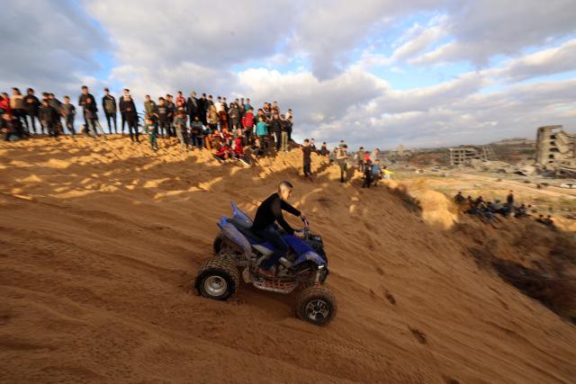 Displaced Palestinians watch as another rides a quad bike on the sand dunes opposite displacement shelters in Nuseirat camp in the central Gaza Strip on December 19, 2025. A famine declared in Gaza in August is now over thanks to improved access for humanitarian aid, the United Nations said on December 19, but warned the food situation in the Palestinian territory remained dire. More than 70 percent of the population is living in makeshift shelters, it said, with hunger exacerbated by winter floods and an increasing risk of hypothermia as temperatures plummet. Although a ceasefire between Israel and militant group Hamas that took effect in October has partially eased restrictions on goods and aid, delivery fluctuates daily and is limited and uneven across the territory, it said. (Photo by Eyad Baba / AFP)