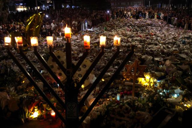 TOPSHOT - A menorah is pictured after the seventh candle is lit for the Jewish festival of Hanukkah as mourners stand next to floral tributes laid out in memory of the victims of a shooting at Bondi Beach, in Sydney on December 20, 2025. Father-and-son gunmen are accused of firing into crowds at a beachside Jewish festival on December 14, killing 15 in an attack authorities linked to "Islamic State ideology". (Photo by DAVID GRAY / AFP)