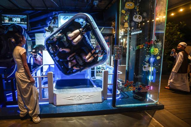 An attendant keeps watch as patrons ride a virtual roller coaster in a simulation booth at an arcade shop in Bangkok on December 20, 2025. (Photo by Candida NG / AFP)