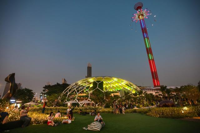 People sit on the lawn in front of the Hatch Dome as passengers take the SkyFlyers giant swing ride in Bangkok on December 20, 2025. (Photo by Candida NG / AFP)