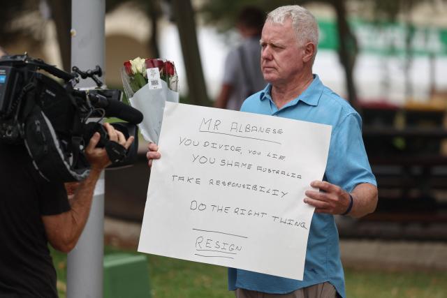 A mourner shows to a TV camera his banner calling for the resignation of Australia's Prime Minister Anthony Albanese as he arrives to attend a memorial held for the victims of a shooting at Bondi Beach in Sydney on December 21, 2025. A father and his son are accused of spraying bullets into the family-thronged Hanukkah celebration at Sydney's most famous beach on December 14, allegedly inspired by "Islamic State ideology". (Photo by DAVID GRAY / AFP)