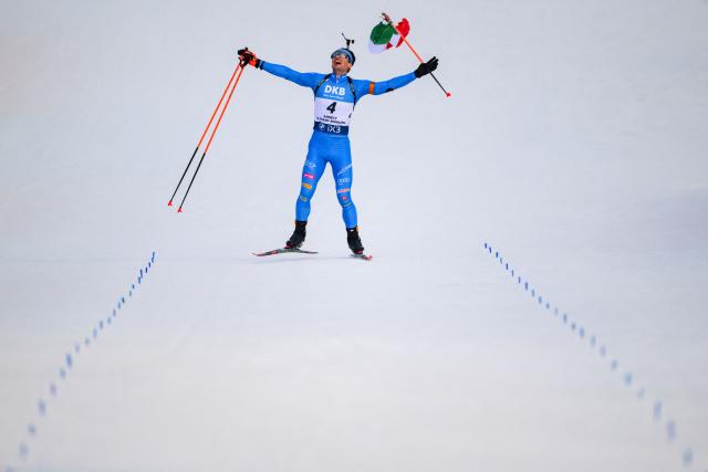 TOPSHOT - Italy's Tommaso Giacomel skies to the finish line with an Italian flag celebrating his win during the men's 15km mass start event of the IBU Biathlon World Cup, in Le Grand Bornand, near Annecy, southeastern France, on December 21, 2025. (Photo by Olivier CHASSIGNOLE / AFP)