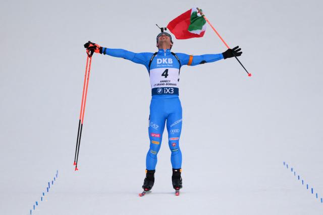 Italy's Tommaso Giacomel skies to the finish line with an Italian flag celebrating his win during the men's 15km mass start event of the IBU Biathlon World Cup, in Le Grand Bornand, near Annecy, southeastern France, on December 21, 2025. (Photo by Olivier CHASSIGNOLE / AFP)