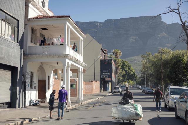 A resident of an occupied building sits on a balcony as a garbage picker rolls down the street in downtown Cape Town on December 18, 2025. Nestled between spectacular mountains and the Atlantic Ocean, Cape Town has emerged as a growing tourism destination, topping in 2025 the Telegraph and Time Out magazine's rankings of best cities in the world. 
Activists say the drive to profit from tourism accommodation is pricing locals out of coveted neighbourhoods, including areas near District Six -- a site long associated with the apartheid-era removals that displaced non-white communities to remote, underdeveloped townships. (Photo by GIANLUIGI GUERCIA / AFP)