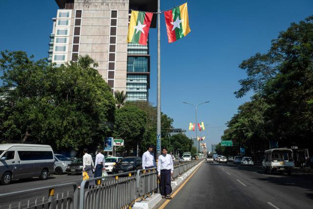 TOPSHOT - Pedestrians wait to cross a road under national flags displayed ahead of Myanmar's general election in Yangon on December 23, 2025. Myanmar junta has touted polls starting December 28 as a path to peace, but the vote will be blocked from rebel-held enclaves and monitors are dismissing it as a ploy to disguise continuing military rule. (Photo by Sai Aung MAIN / AFP)