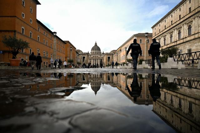 Police officers walk towards the St Peter's Basilica reflected in a puddle along the Via della Conciliazione street in Rome on December 24, 2025. (Photo by TIZIANA FABI / AFP)