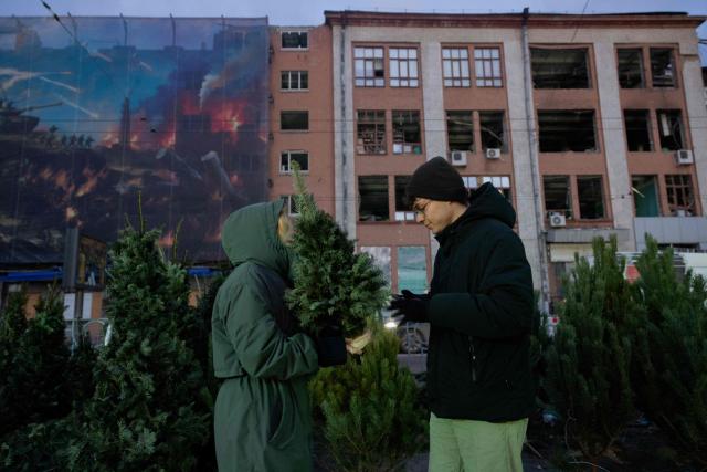 A woman (L) holds a Fir Tree better known as a Christmas tree at a street market near damaged buildings in Kyiv on Christmas Eve celebrations on December 24, 2025, amid the Russian invasion of Ukraine. (Photo by Tetiana DZHAFAROVA / AFP)