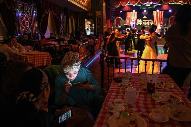 TOPSHOT - This photo taken on December 11, 2025 shows an elderly woman looking at her mobile phone while couples are dancing at the Paramount Ballroom in the Jing'An district of Shanghai. It was just a regular Wednesday at a lunchtime dance club in Shanghai, where dance hall culture has been deeply rooted in the city's identity since its jazz-age heyday. On any given day, multiple venues host hours-long daytime sessions across the finance hub, some starting as early as 6 am. Establishments like the historic Paramount Ballroom are time capsules from the 1930s, while others flash with neon pink and green rave lights in the early afternoon. (Photo by Hector RETAMAL / AFP) / To go with 'CHINA-LIFESTYLE-DANCE-HISTORY-DEMOGRAPHICS, REPORTAGE' by Rita QIAN