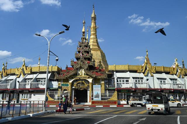 People cross a street in front of the Sule Pagoda in Yangon on December 26, 2025, ahead of the start of Myanmar's general election. Myanmar's junta will preside over elections starting on December 28, advertising the vote as a return to democratic normality five years after staging a coup that triggered civil war. (Photo by Sai Aung MAIN / AFP)