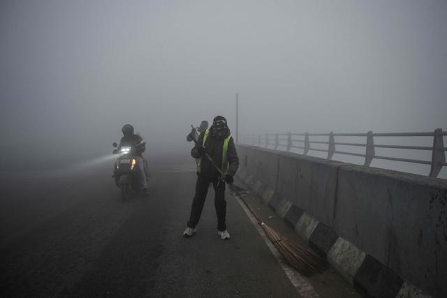 A commuter rides past workers sweeping the Signature Bridge over Yamuna river amid dense smog in New Delhi on December 27, 2025. (Photo by Arun SANKAR / AFP)