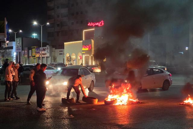 Protesters look on as one place a tire on other burning tires as they protest at a key intersection of the capital with calls to bring down the government, in the Libyan capital Tripoli late on December 26, 2025. (Photo by Mahmud TURKIA / AFP)